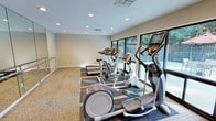 This fitness center is designed to provide a functional and comfortable workout environment. The space is finished with neutral, warm-toned walls and patterned carpet flooring throughout, creating a clean and inviting atmosphere. Recessed lighting is evenly distributed across the ceiling, ensuring the room is well-lit at all times.

Along one wall, a row of large windows spans the length of the cardio equipment area, allowing natural light to fill the space while offering views of the outdoor surroundings, including greenery and what appears to be a patio or pool area beyond. This connection to the outdoors enhances the overall ambiance during workouts.

The cardio equipment lineup includes multiple elliptical trainers, treadmills, and stationary bikes, all positioned to face the windows. On the opposite side of the room, full-length mirrors are mounted along the wall, accompanied by a chrome ballet barre — an ideal feature for stretching, balance exercises, or light barre workouts. This mirrored section visually expands the space, making the room feel larger and more open.

A dedicated open floor area adjacent to the mirrored wall provides additional room for floor exercises or stretching. The space is accessed through a solid white door, offering a private and self-contained fitness environment.
