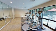 This fitness center is designed to provide a functional and comfortable workout environment. The space is finished with neutral, warm-toned walls and patterned carpet flooring throughout, creating a clean and inviting atmosphere. Recessed lighting is evenly distributed across the ceiling, ensuring the room is well-lit at all times.

Along one wall, a row of large windows spans the length of the cardio equipment area, allowing natural light to fill the space while offering views of the outdoor surroundings, including greenery and what appears to be a patio or pool area beyond. This connection to the outdoors enhances the overall ambiance during workouts.

The cardio equipment lineup includes multiple elliptical trainers, treadmills, and stationary bikes, all positioned to face the windows. On the opposite side of the room, full-length mirrors are mounted along the wall, accompanied by a chrome ballet barre — an ideal feature for stretching, balance exercises, or light barre workouts. This mirrored section visually expands the space, making the room feel larger and more open.

A dedicated open floor area adjacent to the mirrored wall provides additional room for floor exercises or stretching. The space is accessed through a solid white door, offering a private and self-contained fitness environment.