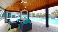 This outdoor amenity space is designed as a fully equipped poolside retreat, centered around a spacious swimming pool with stamped concrete decking throughout. The focal point of the covered cabana is a striking stone fireplace with a glass-front firebox, topped by a wall-mounted flat-screen television — an ideal setup for both relaxing and entertaining. The vaulted cedar-plank ceiling with ceiling fans creates an airy, open-air atmosphere beneath the covered pavilion, while comfortable rattan lounge chairs with teal cushions are arranged for social gathering.

Adjacent to the lounge area, a dedicated outdoor kitchen station features a wide prep counter with a warm wood-panel base and two stainless steel grills, offering a well-appointed space for outdoor cooking. Modern corrugated metal privacy panels line the perimeter, providing both visual separation and a clean, contemporary aesthetic.

Along the poolside, a secondary shaded structure with a translucent teal corrugated roof shelters additional lounge chairs positioned directly beside the water's edge, perfect for relaxing in the shade. Mature trees are visible beyond the enclosure, lending a natural backdrop to this well-designed community outdoor space that seamlessly blends functionality with comfort and style.
