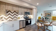 This single-floor residence showcases a thoughtfully designed interior with a cohesive modern aesthetic. The kitchen is a standout feature, combining white lower cabinetry with rustic wood-toned upper cabinets and a bold chevron-patterned tile backsplash. Stainless steel appliances, including a gas range, built-in microwave, dishwasher, and refrigerator, provide a fully equipped cooking space, while wood-look flooring grounds the room with warmth.

The living area offers a comfortable and stylish atmosphere, with neutral walls, wood-look flooring, and a large window that invites natural light and outdoor views. Decorative touches, including a round brass mirror, framed artwork, and a globe floor lamp, contribute to the room's mid-century modern character.

The bedroom continues this aesthetic with a large tufted upholstered headboard, a ceiling fan, and generous windows with outdoor views, lending the space an airy, restful quality.

The bathroom features a soaking tub with a sliding glass enclosure, wood-toned vanity cabinetry with a undermount sink and large mirror, and wood-look tile flooring, creating a clean and functional space. Throughout the residence, consistent flooring and a neutral color palette unify each room into a polished, cohesive whole.