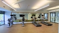 This fitness center is designed to provide a functional and motivating workout environment. The space is anchored by a large, wood-framed wall-to-wall mirror that spans the length of one side, visually expanding the room and allowing users to monitor their form during exercise. The flooring consists of warm hardwood-style material, offering both durability and a clean, polished aesthetic.

The cardio equipment includes multiple treadmills, each equipped with individual monitor screens, and a stationary bike with an attached display. A wall-mounted flat-screen television is also present, providing entertainment options during workouts. The equipment is thoughtfully arranged to maximize floor space and ease of movement between machines.

Overhead, linear fluorescent lighting runs across the ceiling, ensuring the space is evenly and brightly illuminated. The ceiling features exposed structural elements with a clean white finish, contributing to an open, airy feel. A green accent wall, visible in the mirror's reflection, adds a subtle pop of color to the otherwise neutral palette, enhancing the overall energetic atmosphere of the fitness center.