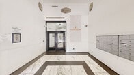 The lobby of the Electric Building Historic Apartments serves as a refined introduction to this historic Fort Worth property, established in 1928. The space features a striking marble-look tile floor laid in a bold geometric border pattern, contrasting elegantly with the dark surrounding tile to create a visually commanding entryway. Crisp white walls are adorned with ornate decorative medallions in the upper corners, nodding to the building's storied architectural heritage. A prominent marble-paneled accent wall displays the Electric Building Historic Apartments branding in gold lettering, reinforcing the property's identity and character. Large glass-paneled entry doors with black frames allow natural light to filter in from W 7th Street, maintaining a bright and welcoming atmosphere. A leasing office with glass storefront is accessible directly from the lobby, providing convenience for residents and prospective tenants. An extensive bank of individual resident mailboxes is neatly recessed into the wall, offering practical functionality. Security cameras are discreetly positioned throughout. The overall ambiance blends historic charm with contemporary updates, making this lobby a polished and functional gateway to one of downtown Fort Worth's notable historic residential addresses.