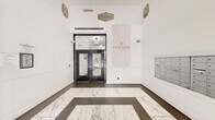 The lobby of the Electric Building Historic Apartments serves as a refined introduction to this historic Fort Worth property, established in 1928. The space features a striking marble-look tile floor laid in a bold geometric border pattern, contrasting elegantly with the dark surrounding tile to create a visually commanding entryway. Crisp white walls are adorned with ornate decorative medallions in the upper corners, nodding to the building's storied architectural heritage. A prominent marble-paneled accent wall displays the Electric Building Historic Apartments branding in gold lettering, reinforcing the property's identity and character. Large glass-paneled entry doors with black frames allow natural light to filter in from W 7th Street, maintaining a bright and welcoming atmosphere. A leasing office with glass storefront is accessible directly from the lobby, providing convenience for residents and prospective tenants. An extensive bank of individual resident mailboxes is neatly recessed into the wall, offering practical functionality. Security cameras are discreetly positioned throughout. The overall ambiance blends historic charm with contemporary updates, making this lobby a polished and functional gateway to one of downtown Fort Worth's notable historic residential addresses.
