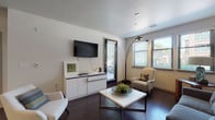 This thoughtfully designed single-floor residence offers a seamless open-concept layout that connects the living, dining, and kitchen areas into one cohesive and functional space. The living area features rich dark hardwood flooring, a contemporary gray sofa accented with patterned throw pillows, and bold abstract artwork adorning the neutral-toned walls. A wall-mounted flat-screen TV, sleek white media console, and direct access to a private outdoor space round out the versatile living zone, which is brightened by natural light through slatted window blinds. The dining area transitions naturally into a well-equipped kitchen, which boasts dark espresso cabinetry, granite countertops, stainless steel appliances including a refrigerator with ice maker and a microwave, and a peninsula with modern bar seating—ideal for casual dining or entertaining. The bedroom is carpeted and serene, featuring a large framed mirror above a white low-profile dresser and easy access to the kitchen and living areas. The bathroom offers a clean, modern aesthetic with large-format tile flooring, dark vanity cabinetry, granite countertops, a wide framed mirror, and bright overhead lighting. Together, these spaces create a cohesive, contemporary residence with a refined yet welcoming atmosphere.