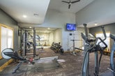 This fitness center is thoughtfully designed to accommodate a comprehensive workout experience across two distinct zones. The main cardio area features soaring ceilings with clerestory windows that flood the space with natural light, complemented by bold burgundy accent walls and gold-trimmed windows that add warmth to the athletic environment. This section houses multiple treadmills, elliptical trainers, and a rowing machine, all positioned on durable, textured carpet flooring with rubber mat underlays for equipment protection. A wooden bench and coat stand provide practical amenities for users.

The strength training zone, separated by sleek glass partitions, features a more subdued color palette with charcoal gray walls and recessed lighting. This area is well-appointed with a power rack, cable machines, a multi-station weight machine, and adjustable benches, catering to a full range of resistance training needs. A wall-mounted flat-screen TV and mirrored walls enhance the functionality of this section. A separate alcove accommodates cycling equipment, rounding out the cardio offerings. The use of gold wood trim throughout ties both zones together cohesively, creating a polished and professional fitness environment.