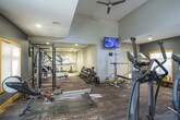 This fitness center is thoughtfully designed to accommodate a comprehensive workout experience across two distinct zones. The main cardio area features soaring ceilings with clerestory windows that flood the space with natural light, complemented by bold burgundy accent walls and gold-trimmed windows that add warmth to the athletic environment. This section houses multiple treadmills, elliptical trainers, and a rowing machine, all positioned on durable, textured carpet flooring with rubber mat underlays for equipment protection. A wooden bench and coat stand provide practical amenities for users.

The strength training zone, separated by sleek glass partitions, features a more subdued color palette with charcoal gray walls and recessed lighting. This area is well-appointed with a power rack, cable machines, a multi-station weight machine, and adjustable benches, catering to a full range of resistance training needs. A wall-mounted flat-screen TV and mirrored walls enhance the functionality of this section. A separate alcove accommodates cycling equipment, rounding out the cardio offerings. The use of gold wood trim throughout ties both zones together cohesively, creating a polished and professional fitness environment.