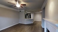 Located in Plano, Texas, this two-story space offers a thoughtfully designed interior that blends comfort and modern style. The main living area showcases rich, dark hardwood flooring that runs throughout, providing a warm and grounded aesthetic against the cool grey walls and crisp white trim and baseboards. Crown molding adds a refined architectural detail that elevates the overall finish of the space.

The open-concept layout connects the living area seamlessly to the kitchen, which features a curved peninsula with a built-in sink, providing both a functional workspace and a natural gathering point. The kitchen is equipped with stainless steel appliances, upper cabinetry, and track lighting that illuminates the space efficiently. A matching set of track lights in the adjacent living area ensures consistent, well-distributed lighting throughout.

A ceiling fan with a built-in light fixture is centrally positioned in the living area, offering both comfort and supplemental lighting. Recessed HVAC vents are integrated into the ceiling, maintaining a clean and uncluttered look. The open sightlines between rooms create an airy, spacious feel, while the two-story configuration provides additional living space and versatility. Overall, the space presents a clean, move-in-ready interior with cohesive design choices throughout.