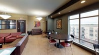 This single-floor communal amenity space is thoughtfully designed to offer a variety of social and recreational environments. The layout encompasses several distinct zones, each with its own character and purpose.

The game room is a standout feature, centered around a full-size pool table set atop a patterned carpet. Large windows flood the area with natural light and offer views of the surrounding urban landscape, while pendant lighting and a cue rack mounted on the wall complete the setup. High-top tables and bar stools provide additional casual seating.

Adjacent to the game room is a comfortable lounge area anchored by a striking fireplace set against a deep red textured accent wall. Rich leather seating arranged around a glass-top coffee table creates an inviting atmosphere for conversation and relaxation.

The bar and kitchenette area features a long dark countertop with red upholstered bar stools, a wall-mounted television, and built-in appliances including a microwave and oven — ideal for entertaining. Warm hardwood flooring and recessed lighting enhance the sophisticated ambiance.

A dining and casual lounge zone offers round tables with red chairs and a sofa seating area, accented by contemporary artwork throughout. The entry lounge near the main door features plush seating and pendant lighting, providing a welcoming transition into the space.
