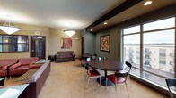 This single-floor communal amenity space is thoughtfully designed to offer a variety of social and recreational environments. The layout encompasses several distinct zones, each with its own character and purpose.

The game room is a standout feature, centered around a full-size pool table set atop a patterned carpet. Large windows flood the area with natural light and offer views of the surrounding urban landscape, while pendant lighting and a cue rack mounted on the wall complete the setup. High-top tables and bar stools provide additional casual seating.

Adjacent to the game room is a comfortable lounge area anchored by a striking fireplace set against a deep red textured accent wall. Rich leather seating arranged around a glass-top coffee table creates an inviting atmosphere for conversation and relaxation.

The bar and kitchenette area features a long dark countertop with red upholstered bar stools, a wall-mounted television, and built-in appliances including a microwave and oven — ideal for entertaining. Warm hardwood flooring and recessed lighting enhance the sophisticated ambiance.

A dining and casual lounge zone offers round tables with red chairs and a sofa seating area, accented by contemporary artwork throughout. The entry lounge near the main door features plush seating and pendant lighting, providing a welcoming transition into the space.