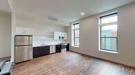 This single-floor unit showcases a contemporary design with an open-concept living and kitchen area anchored by rich, reclaimed-style hardwood flooring. The kitchen is outfitted with dark espresso lower cabinetry contrasted by crisp white upper cabinets and open shelving. A full suite of stainless steel appliances includes a refrigerator, electric range, built-in microwave, and dishwasher, all set against clean white countertops. Tall, floor-to-ceiling windows flood the main living space with natural light while framing striking urban skyline views, reinforcing the elevated city-living aesthetic.

The bedroom features plush neutral carpeting, a generous walk-in closet, and windows offering additional outdoor views. An exposed industrial steel doorframe adds a subtle nod to the building's architectural character. The bathroom is finished with large-format dark tile flooring, a floating dark-wood vanity with a white countertop and integrated sink, and a walk-in glass-enclosed shower fitted with tile surround and a built-in bench. A window in the bathroom provides natural light. Recessed lighting and high ceilings throughout the unit contribute to a bright, spacious ambiance.