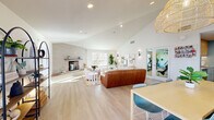 Located at 7425 N Mona Lisa Rd in Tucson, Arizona, this single-floor space offers an open-concept layout that seamlessly integrates a living room and dining area into one cohesive, well-appointed environment. The interior is defined by soaring vaulted ceilings that lend an impressive sense of openness, complemented by crisp white walls and light-toned hardwood-style flooring that runs throughout.

The living area serves as the focal point of the space, anchored by a textured concrete fireplace with a raised brick hearth and a minimalist floating mantle — a striking architectural feature that adds warmth and character. Plush seating, a woven jute area rug, and carefully curated wall art create an inviting, modern bohemian atmosphere. Sleek wall sconces provide accent lighting alongside recessed ceiling fixtures.

The dining area is highlighted by a long natural wood table surrounded by contemporary chairs, set beneath a trio of oversized woven rattan pendant lights that add a warm, artisanal touch. Multiple large windows with clean white blinds flood the space with abundant natural light and offer views of the surrounding outdoor area. A welcoming entry vestibule with a bench and glass French doors provides a graceful transition from the exterior. An arched decorative shelving unit and curated greenery complete the refined, airy aesthetic of this thoughtfully designed space.