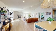 Located at 7425 N Mona Lisa Rd in Tucson, Arizona, this single-floor space offers an open-concept layout that seamlessly integrates a living room and dining area into one cohesive, well-appointed environment. The interior is defined by soaring vaulted ceilings that lend an impressive sense of openness, complemented by crisp white walls and light-toned hardwood-style flooring that runs throughout.

The living area serves as the focal point of the space, anchored by a textured concrete fireplace with a raised brick hearth and a minimalist floating mantle — a striking architectural feature that adds warmth and character. Plush seating, a woven jute area rug, and carefully curated wall art create an inviting, modern bohemian atmosphere. Sleek wall sconces provide accent lighting alongside recessed ceiling fixtures.

The dining area is highlighted by a long natural wood table surrounded by contemporary chairs, set beneath a trio of oversized woven rattan pendant lights that add a warm, artisanal touch. Multiple large windows with clean white blinds flood the space with abundant natural light and offer views of the surrounding outdoor area. A welcoming entry vestibule with a bench and glass French doors provides a graceful transition from the exterior. An arched decorative shelving unit and curated greenery complete the refined, airy aesthetic of this thoughtfully designed space.