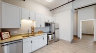 This single-floor unit blends industrial character with modern finishes throughout its open, airy layout. Exposed pipe runs traverse the ceiling, lending a loft-inspired aesthetic that contrasts attractively with the crisp white walls and light wood-look tile flooring found throughout the space. The elevated ceiling height enhances the sense of openness, particularly in the main living and kitchen areas.

The kitchen is well-appointed, featuring white shaker-style cabinetry extending to the upper walls for generous storage, complemented by dark hardware. A full suite of stainless steel appliances includes a refrigerator, range with oven, built-in microwave, and dishwasher. Under-cabinet lighting illuminates the countertops, and a double-basin sink with a modern faucet adds functional detail. A geometric pendant light fixture accents the space.

A separate room offers a quieter retreat, with a window fitted with blinds and a wall-mounted HVAC unit providing climate control. The bathroom is a notable highlight, finished with large-format marble-look wall tiles surrounding the soaking tub and shower combination, accented by a bold vertical stripe of cobalt blue glass tiles that adds a striking visual element. Natural light filters in through a window, completing the bright, clean atmosphere of the space.