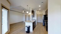This single-floor space showcases an open-concept layout that seamlessly connects the kitchen and living areas. The kitchen is equipped with white shaker-style cabinetry, granite countertops, and a peninsula island with an integrated sink and stainless steel dishwasher. Pendant lighting hangs above the island, adding a refined touch to the functional workspace. The living area extends naturally from the kitchen, benefiting from a ceiling fan and access to natural light through a door with a glass panel. Crown molding runs along the upper perimeter of the walls, lending a clean, finished aesthetic throughout. Durable hardwood-style vinyl plank flooring in a warm gray tone flows continuously across the entire space, providing both visual cohesion and easy maintenance. Recessed lighting and visible HVAC vents are integrated into the white ceiling, ensuring comfort and practical utility. A doorway visible from the main living space suggests additional rooms beyond, while a secondary entry door near the kitchen adds convenient accessibility to the overall floor plan.