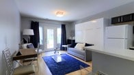 This thoughtfully designed single-floor space showcases a clean, modern aesthetic with a highly functional open-concept layout. The main living area is anchored by warm hardwood flooring and painted in a crisp, neutral gray palette that lends the space a bright and airy feel. A flush-mount crystal chandelier adds a touch of elegance overhead, while a bold navy blue area rug grounds the seating area and introduces a rich pop of color.

A gray sofa with plush accent pillows sits at the heart of the living space, complemented by an acrylic coffee table that keeps the aesthetic light and uncluttered. One of the standout features is the seamlessly integrated white Murphy bed, which folds flush against the wall when not in use, demonstrating smart space utilization without sacrificing style.

Floor-to-ceiling navy drapes frame a set of French doors that open to an outdoor area, allowing natural light to pour in and creating a welcoming indoor-outdoor connection. A dining area with a wood-toned table and modern chairs occupies one side of the room, making the space well-suited for both relaxing and entertaining. Every element of this space has been curated to blend practicality with contemporary design.