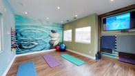 This single-level wellness studio is thoughtfully designed to foster a calm and focused environment for yoga and fitness practice. The room's most striking feature is a large, vivid wall mural depicting balanced zen stones over rippling water, creating an immediately tranquil atmosphere upon entry. The adjacent sage green accent wall complements the mural's cool tones, reinforcing the space's meditative aesthetic. Warm hardwood-style flooring runs throughout, providing a clean, supportive surface well-suited for yoga mats and stretching. Multiple yoga mats in purple, teal, and blue are laid out across the floor, indicating the studio's capacity to accommodate several practitioners simultaneously. Exercise balls in red and blue add versatility for varied fitness routines. Two windows along the green wall allow natural light to filter into the space, enhancing the airy, open feel. Recessed ceiling lighting supplements natural illumination, ensuring the room is well-lit regardless of the time of day. A wall-mounted display and storage area on the far right suggest additional resources for guided workouts. Overall, the studio offers a well-appointed, peaceful retreat designed for wellness.