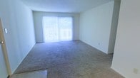 This single-floor space features an open living area finished with wall-to-wall carpeting in a neutral tone, providing a clean and versatile foundation for a variety of furnishing arrangements. The most prominent feature of the room is a large sliding glass door positioned along the far wall, dressed with full-length vertical blinds that allow for adjustable light control. When the blinds are open, the door floods the interior with an abundance of natural light and offers views of the surrounding outdoor greenery visible through the glass. The smooth, white walls throughout the space create a bright and airy atmosphere, complementing the neutral carpeting to give the room a cohesive, understated aesthetic. The layout is straightforward and open, maximizing usable floor space without any unnecessary structural interruptions. The ceiling is finished in a clean, flat style consistent with the overall simplicity of the space. A standard electrical outlet is visible along one wall, and the transition between the carpeted area and a hard-surface flooring section can be seen at the entry point of the room, suggesting a practical separation between living and entry zones. Overall, the space presents a functional and well-lit environment.