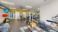 Located at 112 29th Avenue Dr NW in Hickory, NC, this fitness center is a compact yet comprehensively outfitted exercise facility housed on a single floor. The space features a durable rubber flooring surface ideal for high-intensity workouts, and is illuminated by bright fluorescent overhead lighting complemented by natural light streaming through multiple windows. A ceiling fan provides additional air circulation throughout the room.

The strength training area is anchored by a multi-function cable machine and a fully equipped squat/power rack with an adjustable bench, flanked by floor-to-ceiling mirrors that enhance the sense of space and allow users to monitor their form. A dedicated free weights section offers a range of barbells and weight plates.

The cardio zone is outfitted with a treadmill, an elliptical trainer, and a recumbent stationary bike, all positioned to take advantage of the natural light. A water fountain is conveniently mounted on the wall nearby.

Functional training accessories are well-represented, with a rack of medicine balls in various sizes, stability/exercise balls, resistance bands, and foam rollers neatly organized along the walls. The facility's color scheme — crisp white ceilings paired with bold teal and yellow accent walls — creates an energetic and motivating atmosphere. A direct exterior door provides easy outdoor access.