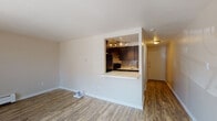 This single-floor residence features a well-organized layout with wood-look flooring running throughout the living areas, creating a cohesive and clean aesthetic. The spacious living room serves as the centerpiece of the unit, highlighted by a striking brick accent wall framing a fireplace and a large sliding glass door that allows natural light to fill the space while providing direct access to the outdoors.

The kitchen is thoughtfully designed with a pass-through opening and a breakfast bar countertop that connects it to the living area. Dark espresso cabinetry is paired with a mosaic tile backsplash, and the kitchen is equipped with a black electric range beneath a ventilation hood with track lighting overhead.

A dedicated entry hallway provides a clear transition into the main living space, while the bathroom features a floating white vanity with a round mirror and hexagonal mosaic tile accent wall. An adjacent reach-in closet with built-in shelving offers practical storage. A baseboard heating system is present in the living area. The overall ambiance is clean and contemporary, with neutral tones creating a versatile backdrop throughout.
