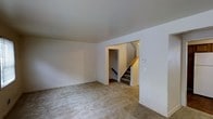 This two-story space features a straightforward, functional layout with neutral finishes that provide a clean, move-in-ready feel. The main level showcases a generously sized open room finished with wall-to-wall carpet in a neutral beige tone, creating a comfortable and versatile living area. The walls are painted in a crisp white, contributing to a bright and airy atmosphere. A centered doorway leads to a dedicated stairwell that connects both floors, framed by white trim and featuring a wooden handrail along carpeted stairs. The stairwell is well-lit, ensuring easy and safe transitions between levels. Electrical outlets and a wall-mounted control panel are thoughtfully positioned throughout the main room, adding to the space's practicality. The smooth, white ceilings maintain a uniform height, reinforcing the open feel of the room. Overall, the space presents a clean and neutral aesthetic, well-suited for a variety of uses and easily adaptable to different furnishing styles.