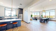 Located at 3500 Cox Rd in Henrico, Virginia, this thoughtfully designed communal space offers a seamless blend of modern interiors and exceptional outdoor amenities. The open-concept layout connects a fully equipped kitchen to a spacious living and dining area, all unified by striking herringbone-patterned light wood flooring. The kitchen features navy cabinetry with gold hardware, white quartz countertops, a large center island with bar seating, stainless steel appliances, and abundant natural light through black-framed windows. The living area flows into a sunlit lounge with floor-to-ceiling windows and a retractable glass garage door that opens to the outdoors, inviting fresh air and panoramic views. A large dining table anchored by sculptural rattan pendant lights provides ample seating for group gatherings. Outdoor spaces include a rooftop terrace equipped with multiple grills, a fire pit dining table, and string lighting, as well as a ground-level patio featuring lounge seating, a fire pit table, and a bar. Together, these spaces create a versatile, welcoming environment ideal for both casual socializing and larger events.