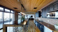 This single-floor space showcases a striking industrial aesthetic through a thoughtful combination of raw materials and modern design elements. Exposed brick walls run throughout, providing a warm, textured backdrop that contrasts beautifully with the natural wood plank ceiling overhead. Wide-plank hardwood floors extend across the open-concept layout, unifying the kitchen, dining, and living areas into one cohesive environment.

A large, dark-stained communal bar table serves as a central focal point, accompanied by metal-frame bar stools and anchored above by an eye-catching industrial pendant light fixture with Edison-style bulbs suspended from black iron piping. The kitchen area features navy-toned cabinetry with dark countertops, a farmhouse-style sink, and a range hood that complements the overall color palette.

Large windows and glass-paneled doors allow natural light to fill the space, enhancing the warmth of the wood tones. The adjoining living area offers a relaxed seating arrangement centered around a wall-mounted television. The overall ambiance is rustic yet refined, combining industrial character with functional, open living.