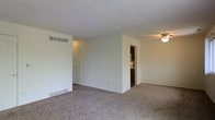 This single-floor residence presents an open and functional layout centered around a spacious living area finished with neutral-toned wall-to-wall carpeting. The room is characterized by clean white walls and a textured ceiling, contributing to a bright, understated aesthetic. A ceiling fan with a light fixture is positioned overhead, providing both air circulation and ambient lighting to the space.

A partial wall divides the living area, creating a subtle sense of separation while maintaining an open feel throughout. Along the base of this dividing wall, a floor-level heating vent is visible, indicating a forced-air climate system. A transom-style window above a doorway allows additional light to pass between rooms, enhancing the overall brightness of the interior.

From the living area, a doorway provides a direct sightline into the kitchen, where dark cabinetry and appliances are visible, suggesting a practical galley-style or compact kitchen configuration. An additional doorway leads to further rooms within the residence. The overall ambiance is clean, neutral, and move-in ready, offering a blank canvas well-suited for a variety of interior arrangements.