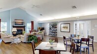 Located at 4111 Wincove Dr in Columbus, Ohio, this single-story community space offers a thoughtfully designed, open-plan interior ideal for gathering and shared activities. The main hall is defined by soaring vaulted ceilings that peak along the central axis, finished in soft blue-gray tones that lend an airy, welcoming atmosphere throughout. Warm hardwood-style flooring runs continuously across the entire space, unifying its distinct functional zones.

The dining area accommodates multiple tables and chairs arranged to encourage group dining, with natural light streaming in through large windows and French doors fitted with an elegant arched transom. A living lounge area anchors one end of the hall, centered around a fireplace with a classic surround, flanked by comfortable seating and a flat-screen television. Decorative open shelving, framed artwork, and curated greenery add character to the space.

A separate lounge nook features additional seating and access through French doors to an adjoining area. The fully equipped kitchen includes warm oak cabinetry, ample counter space, a full range, microwave, and double sink — well-suited for meal preparation. Restroom facilities are also accessible within the space. Recessed and cove lighting throughout ensures a comfortable ambiance at any time of day.