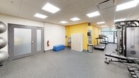 This fitness center is a thoughtfully designed, single-floor facility offering a comprehensive range of exercise equipment and amenities. The space is finished with durable speckled rubber flooring throughout, providing both safety and practicality for high-intensity workouts. A suspended drop ceiling with large recessed LED panel lights ensures the room is consistently and evenly illuminated.

The layout is efficiently divided into dedicated training zones. The cardio area features multiple treadmills positioned near a window, allowing for natural light during workouts. Adjacent to this is a strength training section equipped with a multi-functional cable machine, a flat bench, and a full rack of dumbbells in graduated sizes. Stability balls are neatly stored on a tiered rack, and exercise mats are available for floor-based training.

A striking nature-themed wall mural — depicting a serene autumn forest stream — adds a vibrant, energizing visual element to the space. Accent walls painted in bold yellow further enhance the lively atmosphere. Wall-mounted flat-screen televisions are positioned for easy viewing throughout the facility.

Practical amenities include a water fountain, storage lockers, a seating bench, and a waste receptacle. The facility also contains a separate door — likely leading to a restroom or utility room — as well as a fire extinguisher for safety compliance. Overall, the space is clean, functional, and well-organized.