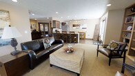 This single-floor space is designed around an open-concept layout that seamlessly connects the living, dining, and kitchen areas into one cohesive environment. The living area anchors the space with a dark leather sofa and a woven ottoman coffee table set atop a neutral carpet, creating a comfortable and grounded focal point. Recessed lighting is distributed evenly across the ceiling, supplemented by a track lighting fixture above the kitchen zone that adds directional illumination and a contemporary touch.

The kitchen features dark espresso cabinetry paired with a stainless steel refrigerator, and a dark-toned island with bar seating for casual dining. The kitchen flows naturally into a dining area with wooden bar-height chairs and a small bistro-style table, reinforcing the informal and functional character of the space. Flooring transitions from carpet in the living area to hard surface in the kitchen and dining zones, visually defining each space without the use of walls.

A secondary room visible through double glass-paneled doors hints at additional functional space, while a window along the far wall introduces natural light. The overall ambiance is warm and modern with a neutral color palette.