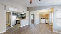 This single-floor residence is designed with an open-concept layout that seamlessly connects the kitchen and living areas, creating a spacious and airy feel. The kitchen features dark cabinetry, stainless steel appliances including a dishwasher and refrigerator, a mosaic tile backsplash, and a peninsula with a sink that opens directly to the main living space — ideal for both cooking and entertaining. Wood-style plank flooring runs throughout the main areas, adding warmth and continuity to the design. A textured ceiling and ceiling fan are present overhead, contributing to the room's functional character. Adjacent to the main living area, a dedicated laundry alcove houses a full-size washer and dryer beneath a pendant light fixture, adding notable convenience. A doorway leads to an additional room with carpeted flooring and a window that allows natural light to filter in. The neutral gray-toned walls and clean white trim provide a modern, versatile backdrop throughout the space, making it well-suited for a variety of décor styles.