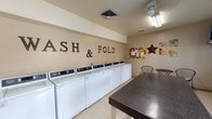 This shared laundry facility is thoughtfully designed to accommodate multiple users simultaneously. The single-level space is outfitted with a generous lineup of front-loading and top-loading washing machines, complemented by both stacked and side-by-side dryer units, providing ample capacity for high-volume use. A bold "Wash & Fold" wall display adds a welcoming, decorative touch to the neutral-toned walls.

A large, dark granite-topped folding table anchors the center of the room, offering substantial workspace for sorting and folding laundry. Adjacent seating with wire bistro chairs and a small table provides a comfortable spot to wait. A utility sink with a standard faucet is conveniently located near the entrance, alongside a trash receptacle and a fire extinguisher for safety compliance.

Natural light filters in through multiple windows, brightening the space alongside overhead fluorescent and pendant lighting. A bulletin board displays community notices, adding a functional, communal element. The flooring is durable and easy to maintain, suited to the demands of a shared facility. The layout flows efficiently, ensuring users can navigate between machines, the folding table, and the utility area with ease.