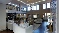 This striking contemporary space is defined by its dramatic double-height ceilings and expansive open-concept layout, creating an immediate sense of grandeur upon entry. The main living area is anchored by a stunning sculptural chandelier — a branching arrangement of globe pendant lights that serves as a bold artistic focal point suspended beneath the soaring ceiling.

The living space is thoughtfully arranged with multiple seating zones, featuring a curated mix of neutral-toned sofas, geometric accent tables, and layered area rugs atop rich dark hardwood flooring. A mezzanine level overlooks the main floor, accessible via an interior walkway framed by sleek metal railings, adding architectural depth and a sense of vertical drama.

Large windows along the perimeter flood the interior with generous natural light, offering views of lush outdoor greenery. Recessed LED lighting and cove lighting accentuate the ceiling lines, complementing the ambient glow throughout. A dedicated office or workspace area with shelving and task chairs is visible in the background, alongside a dining area with an industrial-style table.

The overall ambiance is polished and modern, with a carefully considered neutral palette of whites, grays, and warm wood tones. Wall art and decorative accessories add personality, making this space feel both refined and livable.