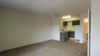 This single-floor residential unit offers a straightforward, functional layout designed for comfortable everyday living. The main living area is finished with wall-to-wall carpeting in a neutral tone, creating a warm and understated atmosphere. A ceiling-mounted light fixture provides overhead illumination, while the textured ceiling adds a subtle layer of visual detail throughout the space.

The living area flows seamlessly into the kitchen, which is partially separated by a raised breakfast bar that doubles as a convenient counter for casual dining or meal prep. The kitchen features dark wood cabinetry that contrasts with the lighter walls and countertops, lending the space a grounded, practical aesthetic. A white refrigerator anchors one end of the kitchen, and under-cabinet lighting brightens the work area, enhancing visibility. The kitchen floor transitions from the carpeted living space to a hard surface, clearly defining each functional zone.

Overall, the space maintains a clean and neutral palette, with beige and off-white tones dominating the walls and surfaces. The open sightlines between the living area and kitchen contribute to a sense of spaciousness, making the unit feel connected and easy to navigate. The layout is well-suited for individuals or small households seeking a practical, move-in-ready living environment.
