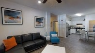 This single-floor residence offers a thoughtfully designed open-concept layout that seamlessly connects the living area and kitchen, creating an inviting and functional space. The living area is anchored by smooth white walls and warm wood-look flooring, lending a clean, modern aesthetic throughout. Recessed lighting and a ceiling fan keep the space well-lit and comfortable. Structural columns subtly define the living zone while maintaining the open feel of the floor plan. The kitchen is outfitted with white cabinetry, stainless steel appliances including a full-size refrigerator, and ample counter space, making it both practical and visually appealing. A pass-through view from the living area to the kitchen enhances the sense of openness. Natural light filters in through window treatments, adding brightness to the interior. The overall ambiance is modern and refreshed, with neutral tones providing a versatile backdrop that suits a variety of décor styles. The thoughtful layout and contemporary finishes make this a well-rounded, move-in ready space suited for everyday living.