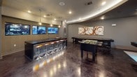 This dynamic entertainment lounge is thoughtfully designed to deliver a premium social and recreational experience. The space is anchored by gleaming, multi-toned polished concrete floors that reflect the recessed and pendant lighting overhead, lending the room a sleek, upscale ambiance throughout.

The game room features a full-sized pool table with a vivid teal felt surface, a foosball table, a shuffleboard table, and multiple electronic dart board arcade machines, as well as a classic Pac-Man tabletop arcade unit — offering a diverse range of entertainment options. Colorful accent chairs and retro-inspired seating are scattered throughout, adding playful pops of orange and teal against the cool grey-toned walls and ceilings.

A bar counter with a dark, textured face and illuminated orange accents runs along one side of the room, lined with sleek chrome barstools. Dual flat-screen televisions mounted above the bar create an ideal gathering spot. The curved ceiling detail in this area adds an architectural flourish that distinguishes the space.

A round dining table with upholstered chairs provides a more relaxed seating zone, while bold artwork — including a large warm-toned landscape painting and abstract canvases — enlivens the walls. Large windows invite natural light, and the open floor plan ensures a comfortable flow between all areas of this well-appointed gathering space.