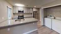 This single-story residence presents a thoughtfully designed layout with a neutral color palette of warm taupe and greige tones throughout. The open-concept living and dining area serves as the heart of the home, highlighted by a white brick fireplace and sliding glass doors that invite natural light and provide outdoor access. Light wood-look flooring flows through the main living spaces, complementing the airy atmosphere enhanced by a ceiling fan overhead.

The kitchen is well-appointed with gray shaker-style cabinetry, granite countertops, and a full suite of stainless steel appliances, including a range, microwave, and refrigerator. A peninsula with an undermount sink adds additional workspace and casual seating potential. A dedicated laundry area with washer and dryer hookups is conveniently situated nearby.

The bedrooms are generously proportioned, each featuring plush carpeting, textured ceilings, and windows that bring in natural light. The bathrooms are finished with granite-topped vanities, gray cabinetry with brushed nickel hardware, and light tile flooring. One bathroom includes a tub-shower combination, while another features a walk-in shower, providing functional variety throughout the home.