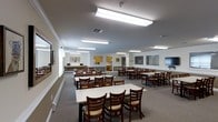 Located at 1000 Nicholas Brooks Ct in New Windsor, New York, this single-floor community space is designed to accommodate group gatherings, dining, and activities with comfort and practicality. The main hall is generously sized, featuring rows of rectangular white tables paired with wooden ladder-back chairs with cushioned seats, offering plentiful seating for a large number of occupants. The room is brightly illuminated by overhead fluorescent lighting panels suspended from a smooth, neutral-toned ceiling, creating a well-lit and welcoming environment throughout. The flooring is covered in a neutral-toned carpet that adds warmth and sound absorption to the space. Along the perimeter, a built-in counter area provides workspace or a service station, while a pass-through window connects to what appears to be a kitchen or service area outfitted with warm wood cabinetry. Wall-mounted monitors and framed artwork add functional and decorative touches to the neutral-painted walls. A decorative wreath near the service area contributes a subtle, welcoming detail. The layout is open and versatile, making it well-suited for communal dining, events, or organized programs.