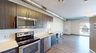 This single-floor unit showcases a contemporary industrial aesthetic, highlighted by exposed cylindrical ductwork that runs prominently through the main living and kitchen areas, adding an urban character to the space. Light wood-tone luxury vinyl plank flooring extends seamlessly throughout, providing a warm, cohesive foundation across every room.

The kitchen is a standout feature, outfitted with dark wood-grain cabinetry reaching toward the ceiling for ample storage, light-toned countertops, and a classic white subway tile backsplash. A full suite of stainless steel appliances includes a refrigerator, electric range, over-the-range microwave, and dishwasher. Track lighting above and a deep single-basin sink complete the functional workspace.

The open living area adjoins the kitchen, offering a generous, flexible layout with clean white walls and natural light from a large window visible in the floorplan view. A dedicated in-unit laundry closet with stacked washer and dryer adds convenience.

The bathroom is cleanly designed with a dark-framed vanity mirror, dark wood-grain cabinetry, white countertop, and accessibility grab bar. The overall ambiance is modern, clean, and functional with consistent industrial-contemporary detailing throughout.