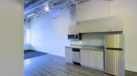 This single-floor space showcases a distinctive industrial-modern aesthetic, blending raw architectural details with contemporary finishes. The open-plan layout offers a generous, flexible living or working area enhanced by soaring ceilings that amplify the sense of space and volume. Exposed ductwork and overhead pipes run along the ceiling, adding an authentic industrial character that contrasts stylishly with the clean white walls below. Durable wood-look flooring extends throughout, providing warmth and cohesion to the open environment. A functional kitchenette lines one wall, featuring light wood cabinetry, a stainless steel sink, a built-in dishwasher, an over-the-range microwave, and a full-sized refrigerator — offering a complete and efficient culinary setup. Track lighting overhead ensures the space is well-lit throughout. Near the entryway, a dark-toned mat provides a practical transition zone. The overall ambiance is clean, airy, and contemporary, making this space well-suited for a variety of uses, from modern residential living to creative or professional work environments.