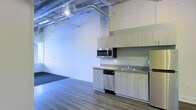This single-floor space showcases a distinctive industrial-modern aesthetic, blending raw architectural details with contemporary finishes. The open-plan layout offers a generous, flexible living or working area enhanced by soaring ceilings that amplify the sense of space and volume. Exposed ductwork and overhead pipes run along the ceiling, adding an authentic industrial character that contrasts stylishly with the clean white walls below. Durable wood-look flooring extends throughout, providing warmth and cohesion to the open environment. A functional kitchenette lines one wall, featuring light wood cabinetry, a stainless steel sink, a built-in dishwasher, an over-the-range microwave, and a full-sized refrigerator — offering a complete and efficient culinary setup. Track lighting overhead ensures the space is well-lit throughout. Near the entryway, a dark-toned mat provides a practical transition zone. The overall ambiance is clean, airy, and contemporary, making this space well-suited for a variety of uses, from modern residential living to creative or professional work environments.
