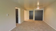 This single-floor space presents a straightforward and functional layout finished in neutral tones. The main living area is carpeted in a warm beige, providing a comfortable underfoot feel that extends throughout the room. Walls are painted in a soft, muted tone that contributes to a clean and understated aesthetic.

A ceiling fan with an integrated light fixture is centrally positioned, offering both air circulation and ambient lighting. The room features multiple dark-stained doors, including what appears to be a double-door closet, offering generous built-in storage. An additional single closet or utility door is visible along the adjacent wall, further enhancing storage options.

The layout includes an open entryway leading to an adjoining area, visible through a doorway in the background, suggesting a connected flow between spaces. White baseboards provide a crisp contrast against the neutral carpet and walls. The overall ambiance is simple and move-in ready, suited for straightforward furnishing and personalization. The space is well-suited for those seeking a functional, no-frills environment with adequate storage and comfortable living space on a single level.