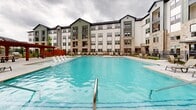 Located at 1540 Edgefield Dr in Garland, Texas, this outdoor amenity space centers around a generously sized swimming pool with crystal-clear blue water, designed to serve as a resort-style retreat for residents. The pool deck is finished in smooth concrete and provides ample space for relaxation, lined with multiple chaise lounges and complemented by several outdoor dining table-and-chair sets ideal for gathering and socializing.

A standout architectural feature of the space is the warm-toned wood pergola structure, which offers shaded seating and an outdoor grilling station, adding both functional and aesthetic character to the area. The surrounding grounds are neatly landscaped with manicured greenery and planting beds, enclosed by a black metal perimeter fence that maintains both security and an open visual connection to the broader community.

The pool area is framed on multiple sides by modern multi-story residential buildings constructed with a mix of stone, board-and-batten siding, and neutral exterior tones, creating an attractive and cohesive architectural backdrop. Tall light poles are positioned throughout the deck to ensure the space remains well-lit and usable into the evening hours. The overall design of this outdoor space prioritizes comfort, social interaction, and a polished community aesthetic.