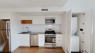 This two-floor space showcases a clean, modern design with a consistent white palette and thoughtful finishes throughout. The kitchen is a standout feature, outfitted with flat-front white cabinetry, dark countertops, a white subway tile backsplash, and a full suite of stainless steel GE appliances including a gas range, built-in microwave, and dishwasher. A warm wood-accented open cabinet adds visual contrast to the otherwise crisp aesthetic. The kitchen also includes an in-unit stacked washer and dryer tucked neatly into an adjacent alcove. Wood-plank flooring runs through the lower level, contributing warmth and continuity to the space.

The upper level offers an open, airy layout with plush carpeting, recessed lighting, and pendant lights that illuminate the space with a soft glow. A glass-panel railing system at the staircase opening enhances the sense of openness while maintaining a sleek, contemporary look. The upper floor's clean white walls and open sightlines create a versatile environment well-suited for various uses.

The bathroom features hexagonal tile flooring in a neutral gray tone, a wood-vanity with an undermount sink and white countertop, a large mirror, and marble-accented wall panels, rounding out the space with refined, modern detailing.