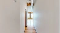 Located at 1 Websters Landing in Syracuse, NY, this thoughtfully designed unit blends historic industrial character with contemporary finishes. The open layout is anchored by rich hardwood floors that run throughout the main living area, complemented by soaring ceilings with exposed original timber beams and visible ductwork that speak to the building's storied past.

A standout feature of the living area is the original brick accent wall, framing a trio of large windows with wooden sills that flood the space with natural light and offer elevated views of the surrounding Syracuse cityscape and tree-lined streets.

The kitchen is well-appointed with white shaker cabinetry reaching ceiling height, granite countertops, a peninsula with a built-in sink, stainless steel appliances including a refrigerator, electric range, and microwave, all illuminated by modern pendant lighting. A sliding barn door adds a functional yet stylish industrial touch in the hallway, which connects the main living areas to the bathroom and two closets.

The bathroom features tile flooring in warm neutral tones, a white vanity with granite countertop, and accessibility-friendly grab bars. Two closets provide ample storage throughout the unit. This space offers a compelling combination of historic architectural details and modern comfort in the heart of Syracuse.