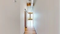 Located at 1 Websters Landing in Syracuse, NY, this thoughtfully designed unit blends historic industrial character with contemporary finishes. The open layout is anchored by rich hardwood floors that run throughout the main living area, complemented by soaring ceilings with exposed original timber beams and visible ductwork that speak to the building's storied past.

A standout feature of the living area is the original brick accent wall, framing a trio of large windows with wooden sills that flood the space with natural light and offer elevated views of the surrounding Syracuse cityscape and tree-lined streets.

The kitchen is well-appointed with white shaker cabinetry reaching ceiling height, granite countertops, a peninsula with a built-in sink, stainless steel appliances including a refrigerator, electric range, and microwave, all illuminated by modern pendant lighting. A sliding barn door adds a functional yet stylish industrial touch in the hallway, which connects the main living areas to the bathroom and two closets.

The bathroom features tile flooring in warm neutral tones, a white vanity with granite countertop, and accessibility-friendly grab bars. Two closets provide ample storage throughout the unit. This space offers a compelling combination of historic architectural details and modern comfort in the heart of Syracuse.