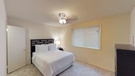 This single-floor space presents a warm and inviting atmosphere defined by its neutral color palette and thoughtful layout. The bedroom is a standout feature, showcasing soft wall-to-wall carpeting in a light beige tone that adds comfort underfoot. Walls are finished in a gentle cream hue, contributing to the room's calm and restful ambiance. A dark-stained wooden bed frame with a classic lattice headboard serves as a focal point, complemented by crisp white bedding and coordinating bedside lighting that enhances the cozy feel. A ceiling fan with an integrated light fixture is centrally mounted, offering both illumination and climate comfort. Natural light filters into the room through a generously sized window dressed with horizontal blinds, allowing for flexible light control and privacy. The overall design is clean and understated, prioritizing comfort and functionality. The consistent use of neutral tones throughout creates a cohesive, serene environment well-suited for rest and relaxation.