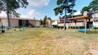 This single-story residence is part of a brick multi-building complex surrounded by mature trees and a spacious shared lawn. The outdoor common area features a pergola, open green space, and a small play area, offering a relaxed communal environment beneath a broad, open sky.

Inside, the interior design is thoughtfully curated, blending raw and refined elements throughout. The floors are finished in dark polished concrete, grounding the space with an industrial character that contrasts elegantly with the warm, neutral-toned walls. Recessed lighting illuminates the rooms from above, complementing the clean, minimalist aesthetic.

The dining area serves as a standout feature, centered around a striking round green marble pedestal table surrounded by woven rattan chairs and anchored by a woven area rug. A large framed artwork with soft, organic textures adorns the wall, adding an artistic focal point. A sliding barn door with a black metal track connects rooms, introducing rustic charm. Adjacent spaces are accessed through weathered vintage-style French doors, adding layers of character. The overall ambiance is eclectic, curated, and inviting.