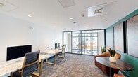 This contemporary office space is thoughtfully designed to balance productivity and comfort across a single floor. The layout is anchored by sleek marble-topped workstations with gold-accented legs and ergonomic black office chairs, providing a polished and professional environment for focused work. Complementing the work area is a cozy lounge corner furnished with curved cognac leather chairs arranged around a round coffee table, ideal for informal meetings or relaxed collaboration.

A striking teal-green accent wall adds a bold pop of color to the interior, contrasting elegantly with the crisp white walls and ceilings throughout. Framed artwork adorns the accent wall, contributing to the space's curated aesthetic. The flooring features patterned, multi-toned carpeting that adds warmth and texture underfoot.

Floor-to-ceiling black-framed glass windows and a glass-panel entry door flood the space with natural light while offering open views of an adjacent outdoor courtyard with seating and vibrant umbrellas. Recessed ceiling lighting and integrated HVAC vents ensure a comfortable and well-lit environment at all times. The glass-paneled door leading to adjacent areas maintains visual connectivity while preserving a sense of defined, purposeful workspace. Overall, the space exudes a modern, upscale office ambiance.