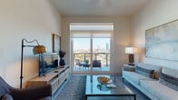 This well-appointed single-floor residence offers a thoughtfully designed layout that blends modern aesthetics with functional living. The open-concept living area is anchored by large sliding glass doors that frame sweeping city skyline views and open directly onto a private balcony, furnished for outdoor relaxation. Soaring ceilings amplify the sense of space, while neutral tones and contemporary furnishings create a clean, inviting ambiance throughout.

The kitchen is a standout feature, showcasing white cabinetry extending to the ceiling, a dark tile backsplash, stainless steel appliances including a full-size refrigerator, dishwasher, and range, as well as a generous center island with bar seating and pendant lighting overhead. Light wood-style flooring ties the kitchen and living areas together seamlessly.

The primary bedroom enjoys direct city views through a large window, offering an airy retreat with plush carpeting. Its en-suite bathroom features a dual-sink vanity with white cabinetry, large-format tile flooring, a walk-in glass shower, and access to a dedicated walk-in closet. A second bedroom provides a comfortable, serene space with soft carpeting and warm lighting. A second full bathroom serves the additional bedroom with matching clean, modern finishes throughout.